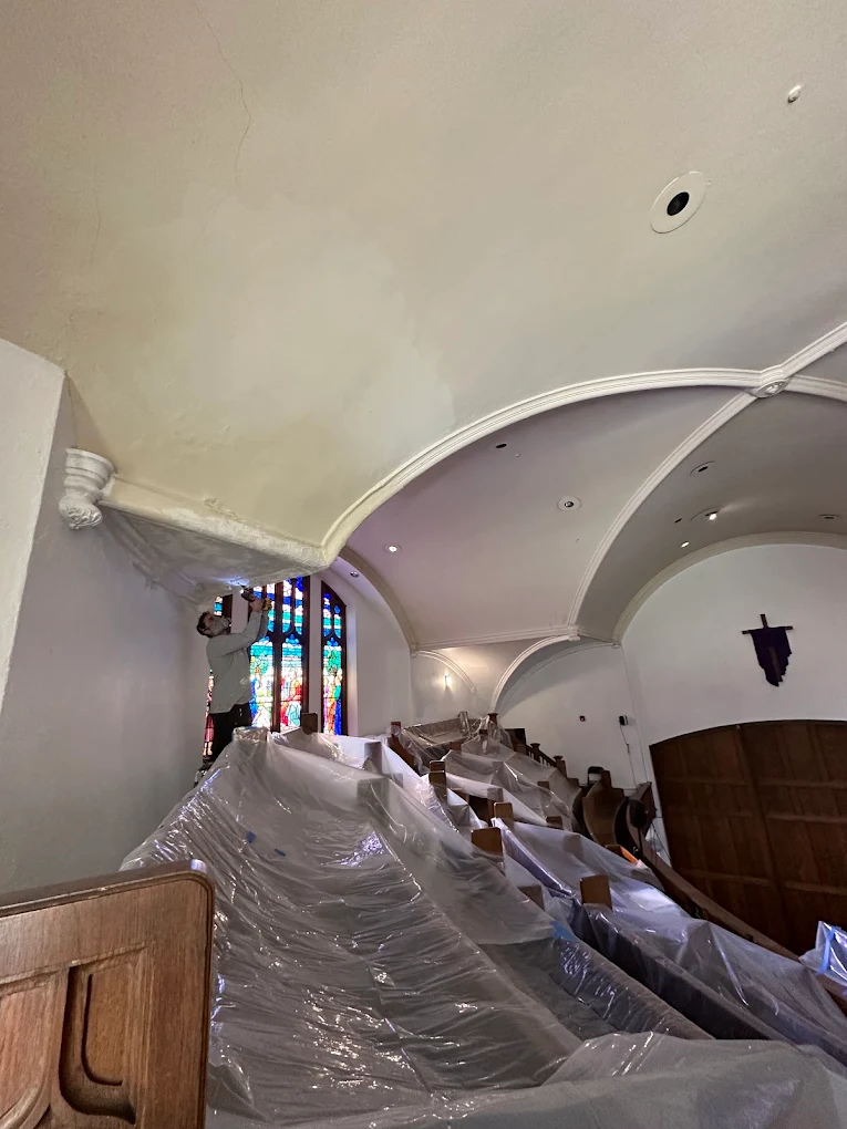 Painter restoring the vaulted ceiling above protected church pews